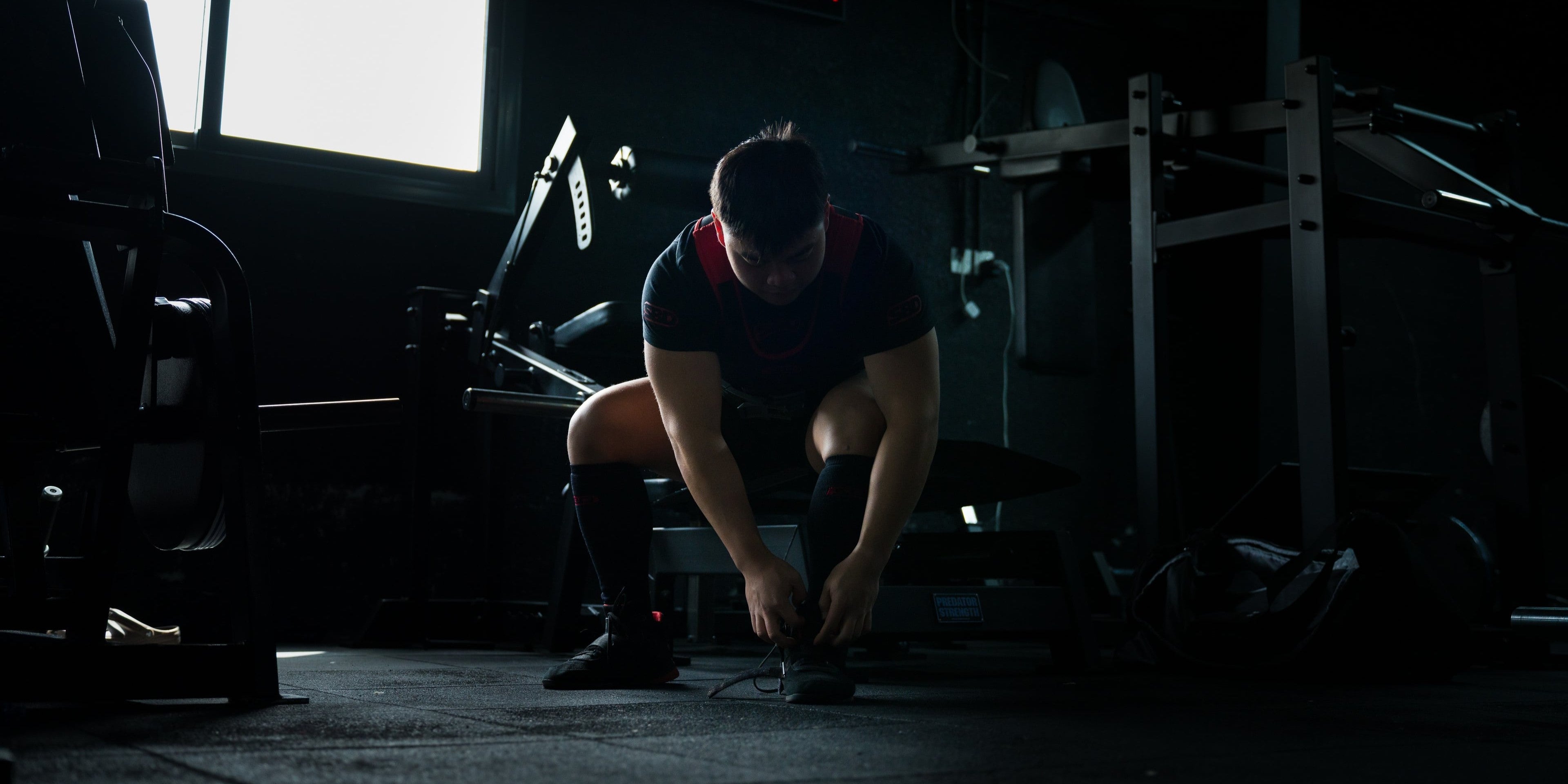 Person wearing a classic SBD competition set tying shoes in a dark gym setting