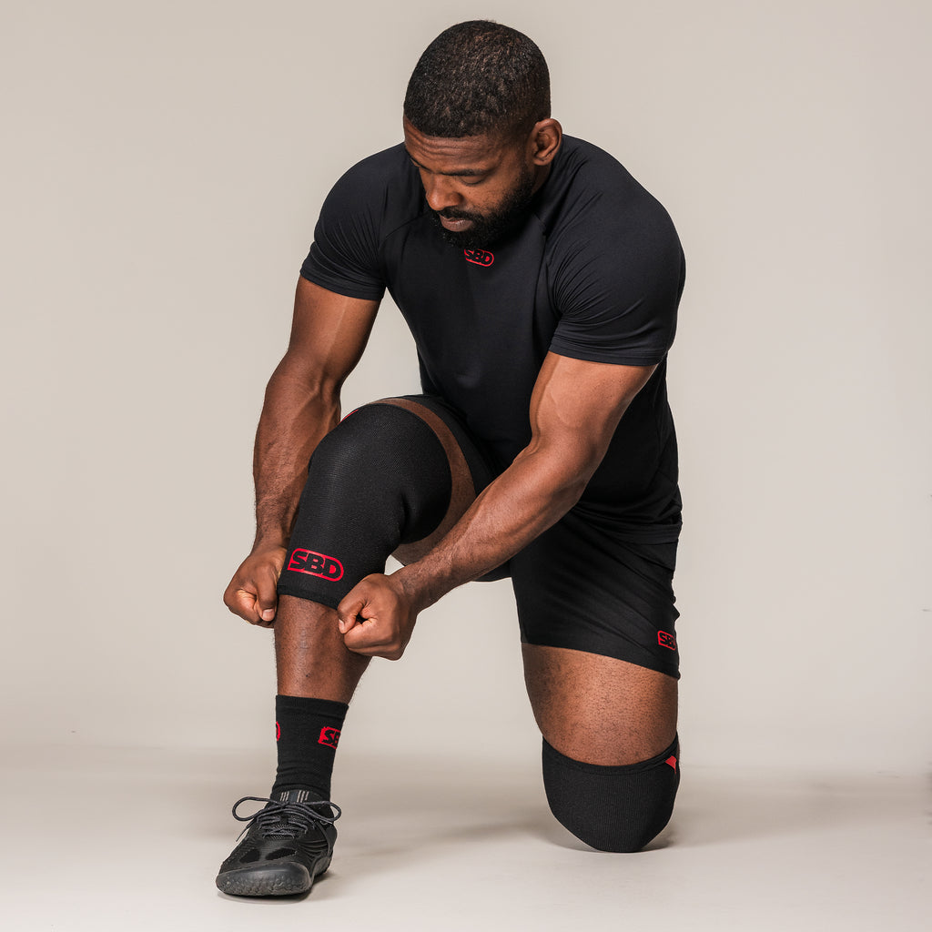 Kneeling athlete adjusting black and red SBD Powerlifting Knee Sleeves