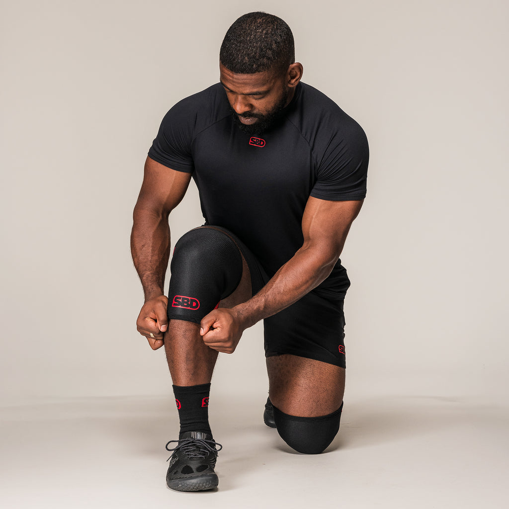 Kneeling athlete adjusting black and red SBD Weightlifting Knee Sleeves